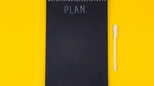 Melna piezīmju grāmatiņa ar uzrakstu Plan uz koša dzeltena fona.