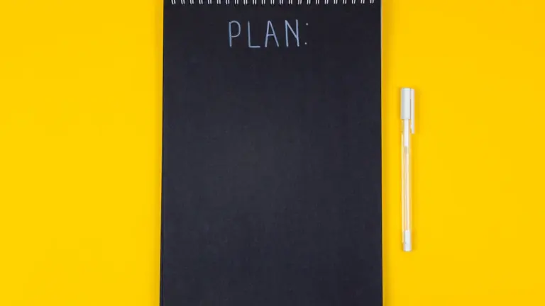 Melna piezīmju grāmatiņa ar uzrakstu Plan uz koša dzeltena fona.