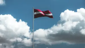 Plīvojošs Latvijas valsts karogs uz augsta masta pretī zilām debesīm ar mākoņiem.