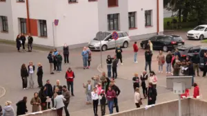 Cilvēku grupa pulcējusies pagalmā pie daudzdzīvokļu ēkas, lai piedalītos āra aktivitātē vai pasākumā.