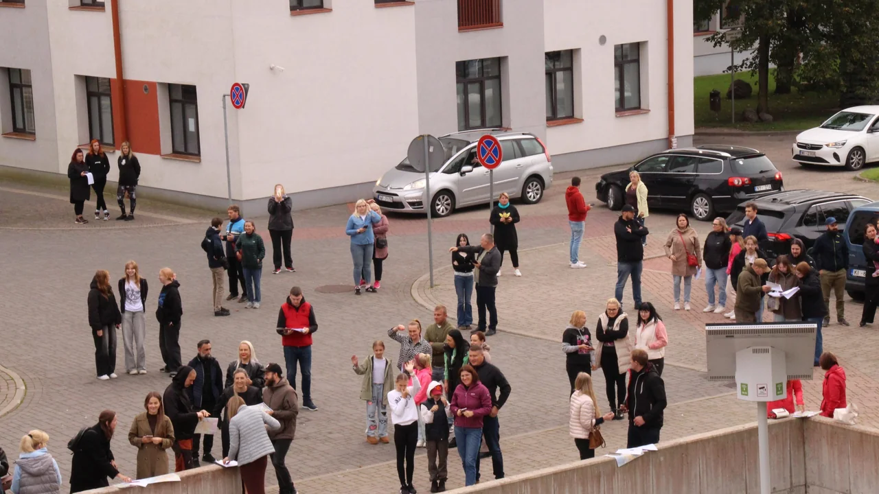 Cilvēku grupa pulcējusies pagalmā pie daudzdzīvokļu ēkas, lai piedalītos āra aktivitātē vai pasākumā.