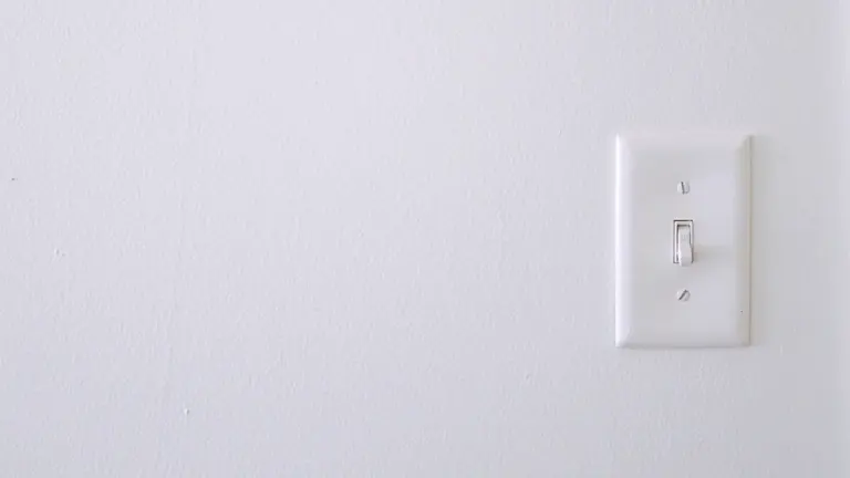 Vienkāršs balts gaismas slēdzis uz gaišas sienas, simbolizējot elektroenerģijas taupīšanu mājās.