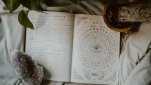 Atvērta grāmata ar astroloģiskiem simboliem un kristālu uz mīksta auduma fona.
