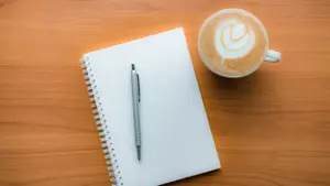 Kafijas tase ar bloknotu un pildspalvu uz koka galda pārdomām par darbu